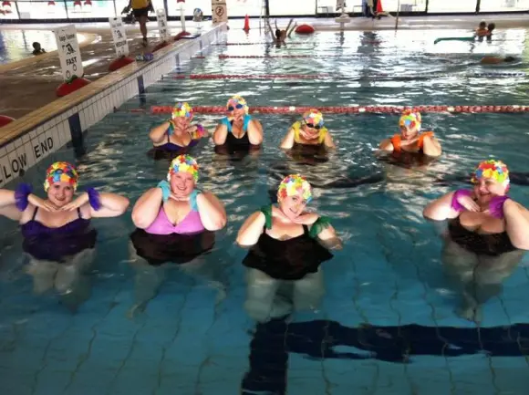 A picture of 8 fat ladies in a swimming pool.  They are all wearing floral swimming caps and posing with their hands underneath their chin.